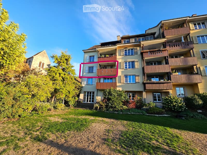 Multi-story residential building with multiple balconies, surrounded by greenery and a lawn.