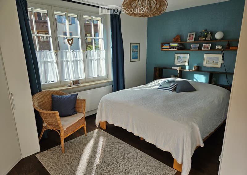 Cozy bedroom with a double bed, wicker chair, shelves with books and decor, and a hanging wicker light fixture.