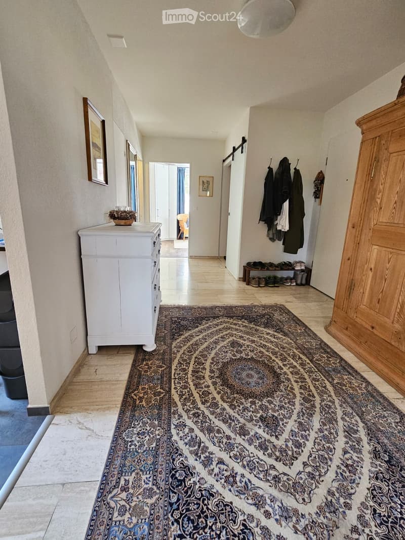 Spacious hallway with beige tiled flooring, large patterned rug, white cabinet, coat rack with jackets, and framed artwork on the walls.
