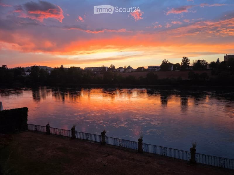 A serene riverside view at sunset, with calm water reflecting the vibrant orange and pink sky, surrounded by trees and distant buildings.