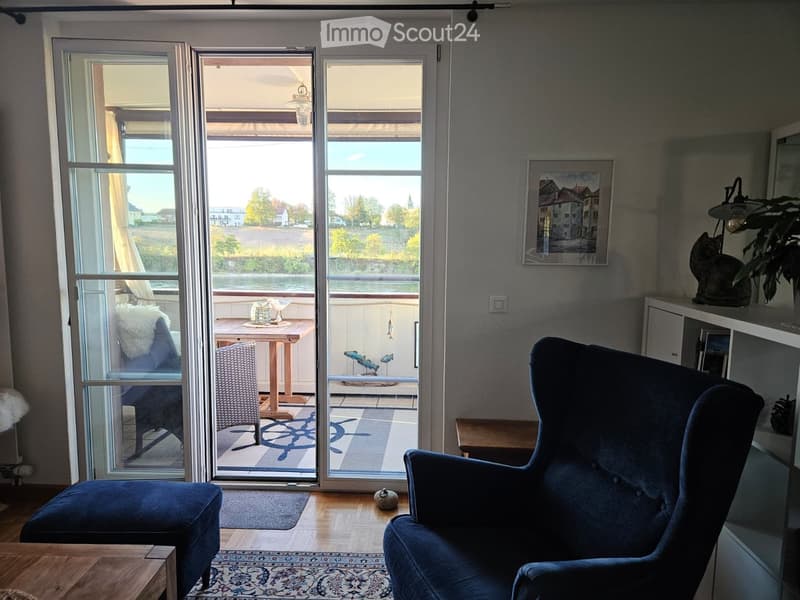 Living room with large windows and a view, featuring a blue sofa, armchair, coffee table, shelf with a picture frame and plant, and a small table on the balcony.