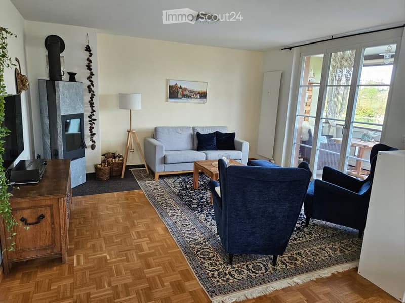 Modern living room with wooden flooring, a fireplace, a sofa, armchairs, a coffee table, and a floor lamp. Large windows and a sliding glass door lead to a balcony.