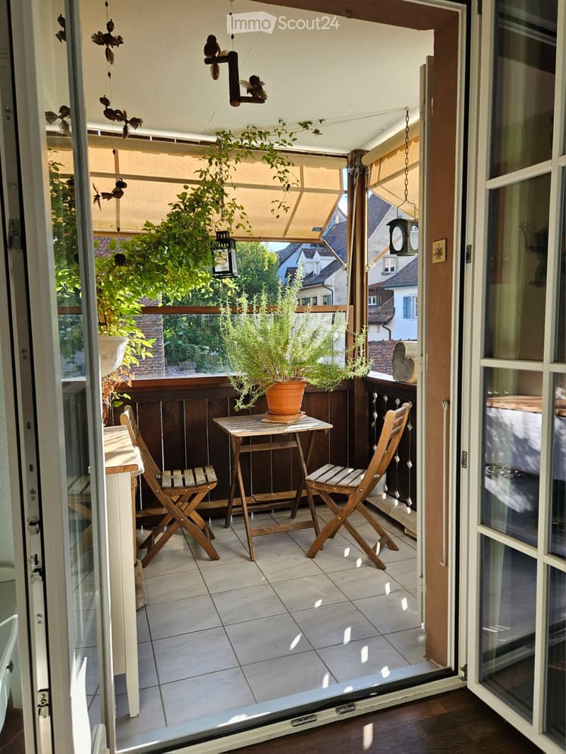 Balcony with tiled floor, small wooden table, two folding chairs, and potted plants. The balcony has a wooden railing and is partially covered by a canopy. The view shows neighboring houses and greenery.