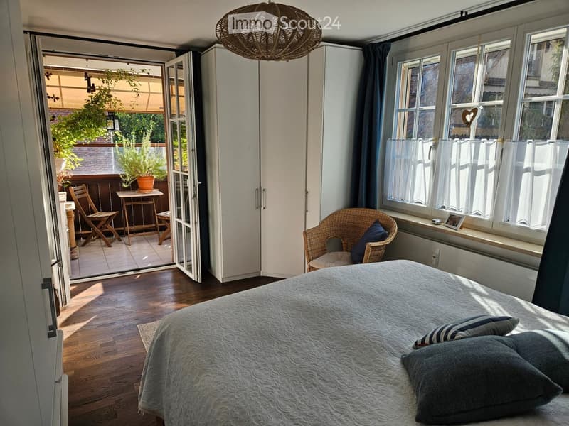 Cozy bedroom with wooden flooring, large windows with white curtains, a bed with grey bedding, a wicker chair, and an open sliding door leading to a small balcony with a table and chairs.