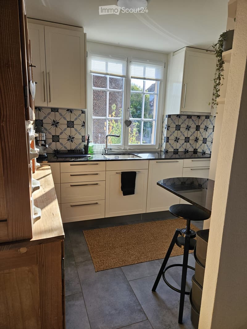 Modern kitchen with light-colored cabinets, a black countertop, and a sink with a modern faucet. A small black stool and a round table are present, along with a coffee maker and a small rug on the floor.
