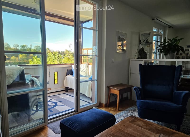Bright living room with large windows and sliding glass doors leading to a balcony. Features a blue sofa, ottoman, wooden coffee table, armchair, side table, lamp, and decorative plant.