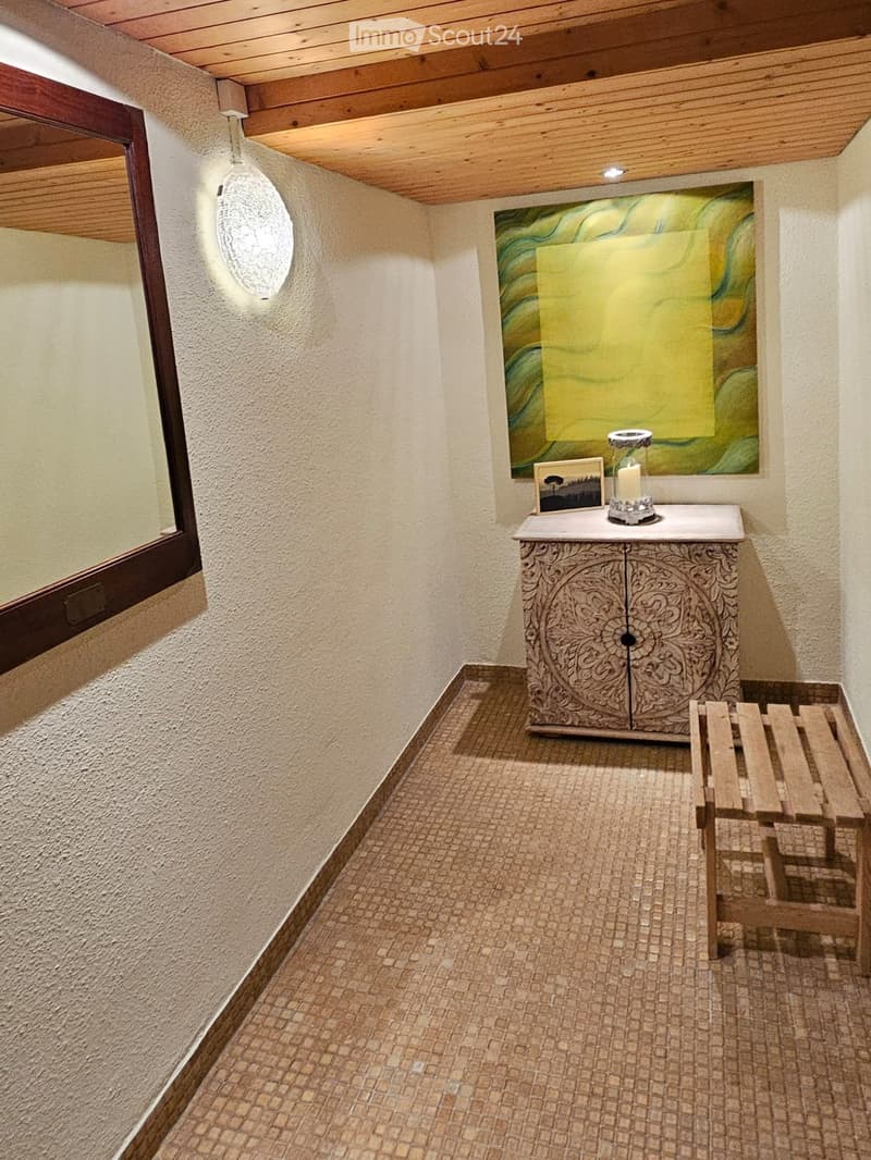 A narrow hallway with textured beige walls, a wooden bench, a small ornate table with a candle, and a modern abstract painting on the wall.