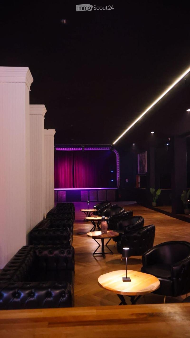 A modern lounge area with dark leather sofas, wooden tables, and ambient lighting, featuring a stage area with curtains in the background.