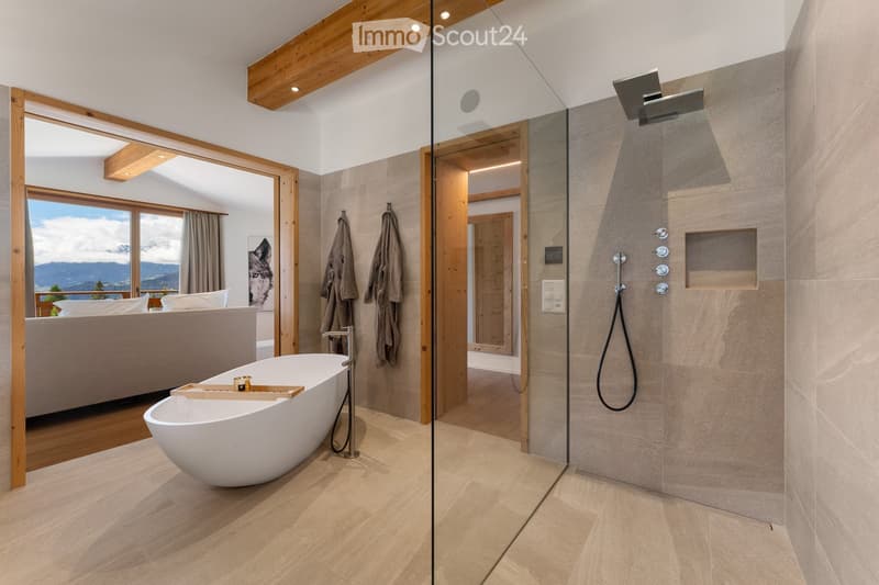 Modern bathroom with a freestanding bathtub, glass-enclosed shower, wooden accents, and a view of a mountain landscape through large windows.