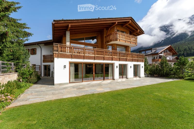 A large, modern two-story house with white walls, wooden balconies, and large windows, surrounded by a well-maintained lawn and trees, with a scenic mountain view in the background.