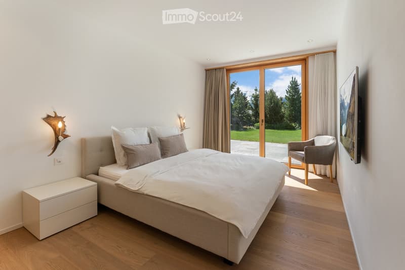 Modern bedroom with a large bed, nightstand, wall-mounted lamp, chair, and a large window with a view of greenery.