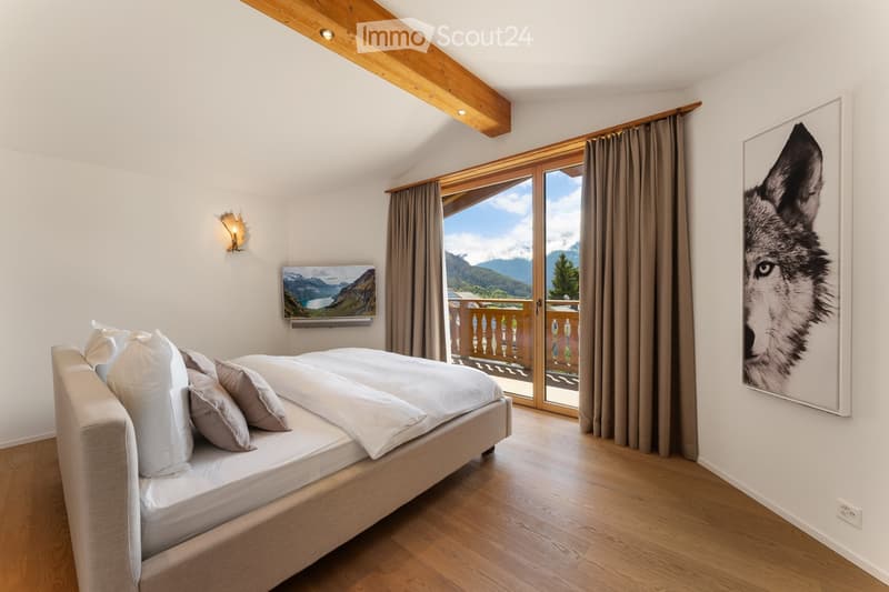 Cozy bedroom with a large bed, pillows, wall-mounted TV, and a scenic mountain view through large windows with curtains.