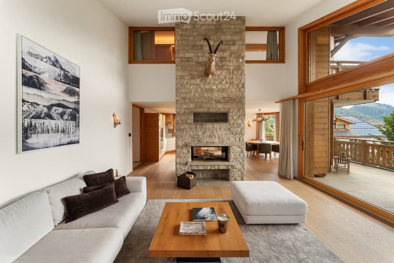 Modern living room with a stone fireplace, large windows offering a scenic mountain view, a white sofa with dark cushions, a wooden coffee table, and an ottoman. A dining area with a round table and chairs is visible in the background.