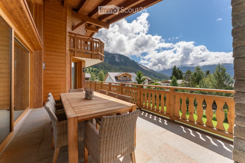 Balcony with wooden table and wicker chairs, overlooking a scenic mountain view.