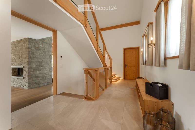 Spacious hallway with wooden flooring, a curved staircase with glass railing, a stone fireplace, and a wooden bench with decorative items.