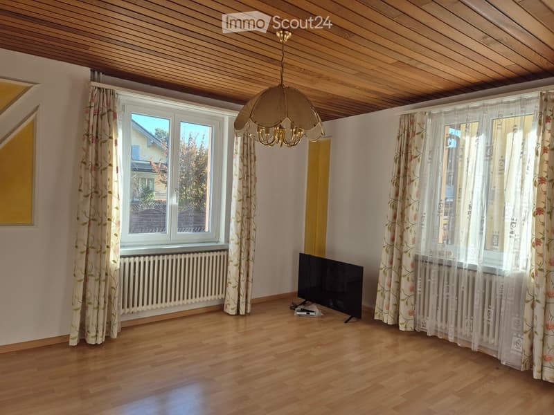 Empty living room with wooden flooring, large windows with floral curtains, a television, radiator, and a hanging chandelier.