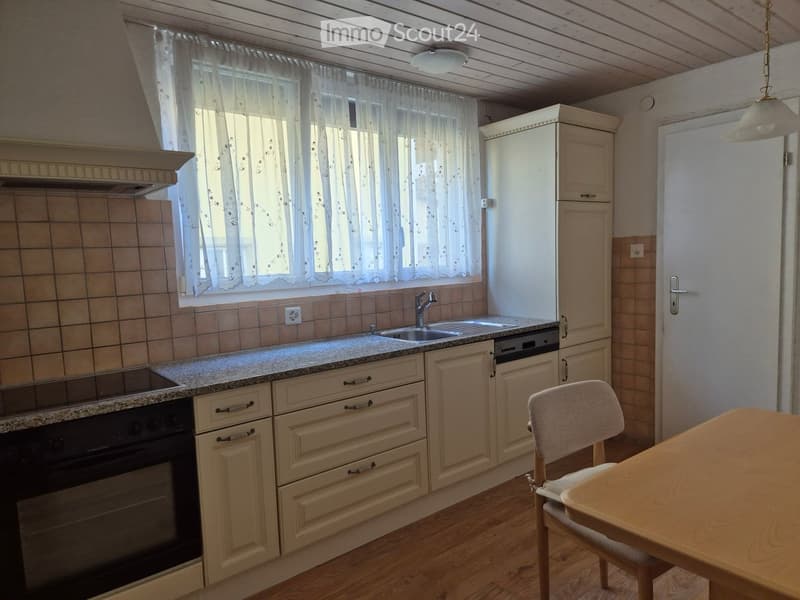 Bright kitchen with light wooden cabinets, granite countertop, stainless steel sink, and appliances including a refrigerator, oven, and dishwasher. A small dining area with a table and chair is visible.
