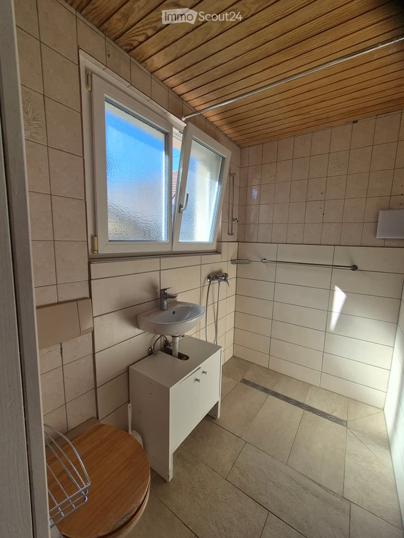 Modern bathroom with wooden ceiling, tiled walls, a sink with a cabinet, and a toilet. Features a window with frosted glass and a shower area with a handheld showerhead.