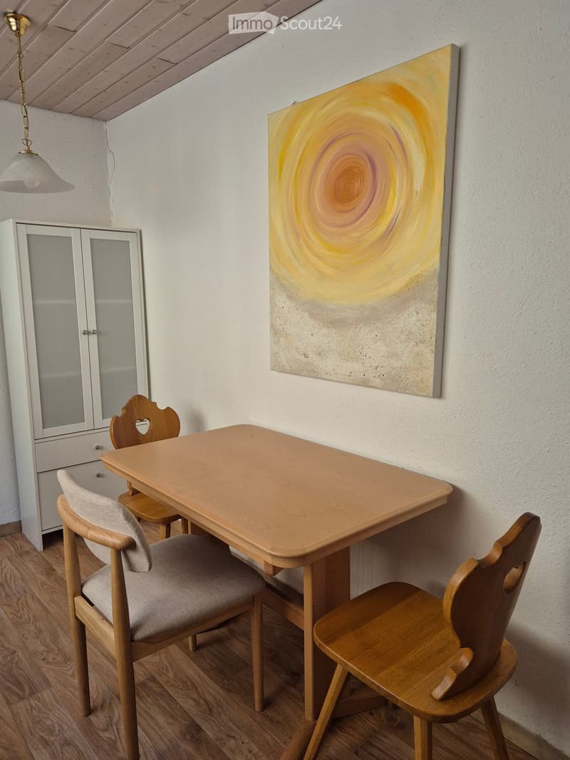 A small, cozy dining area with a wooden table, four chairs, a white cabinet, and a large abstract painting on the wall.