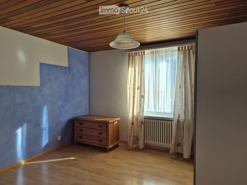 Empty room with wooden floor, blue walls, a window with curtains, a radiator, and a wooden chest of drawers.