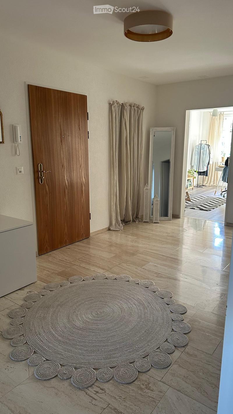 Spacious hallway with tiled flooring, large wooden door, round woven rug, mirror, and beige curtains.