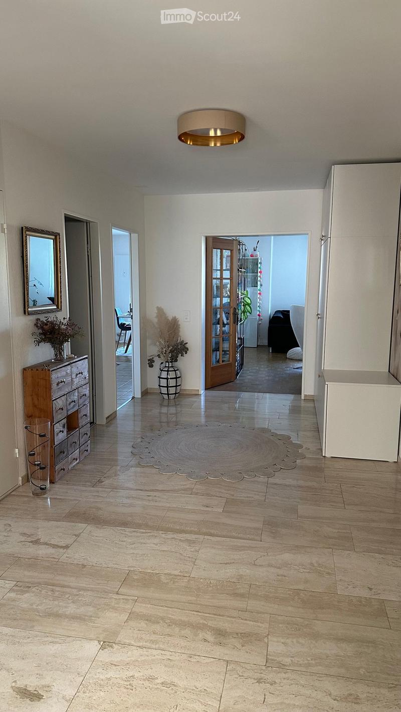 Spacious hallway with polished tile flooring, a central round rug, and several doorways leading to other rooms. Features a wooden dresser with drawers, a mirror, and decorative vases with dried plants.
