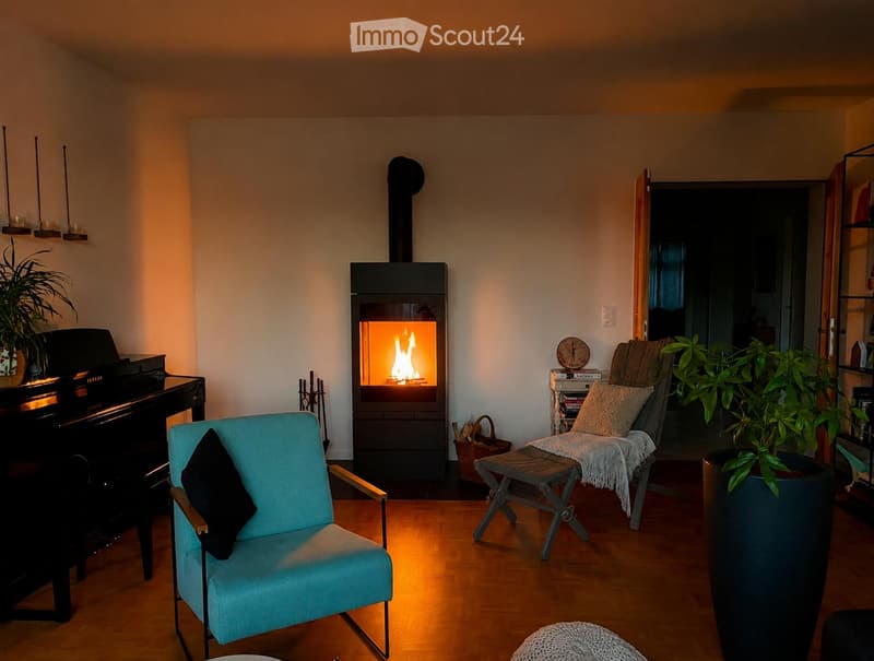 Cozy living room with a modern fireplace, a blue armchair, a small stool, a piano, and a potted plant. The room features warm lighting and wooden flooring.