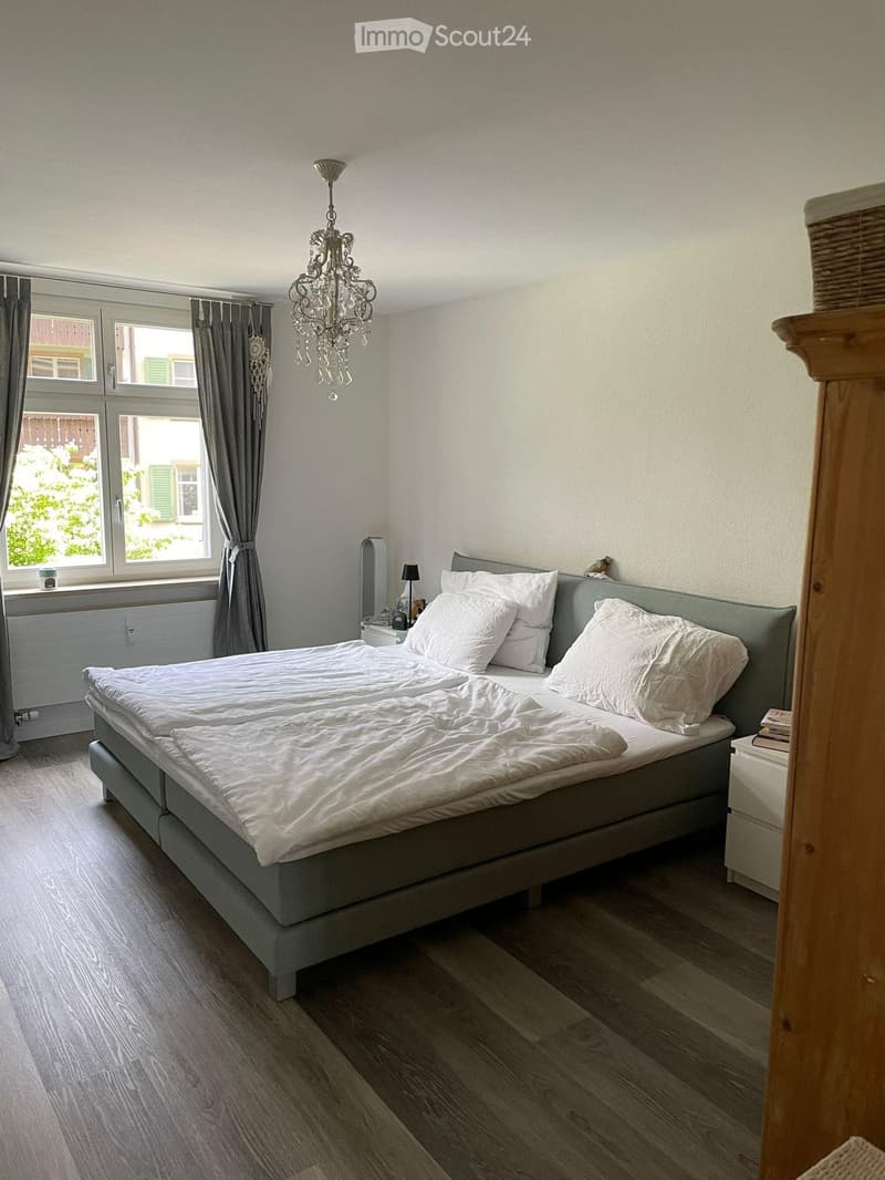 A well-lit bedroom with a large bed, white bedding, a chandelier, and a nightstand with a lamp. The room features wooden flooring and large windows with curtains.