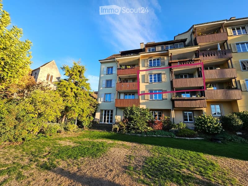 Multi-story residential building with multiple balconies, surrounded by greenery and a lawn.