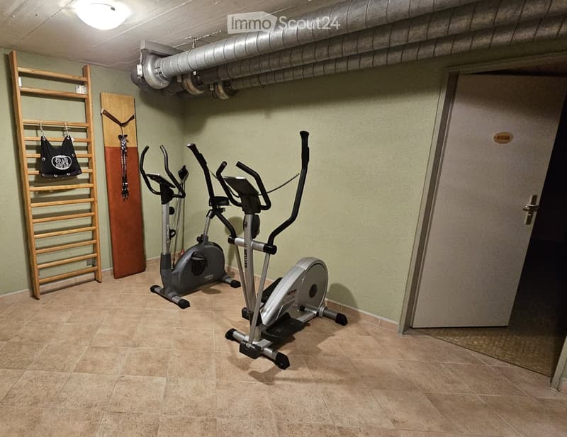 A well-lit basement room with tiled flooring, two elliptical trainers, a wooden ladder, and a clock on the wall.