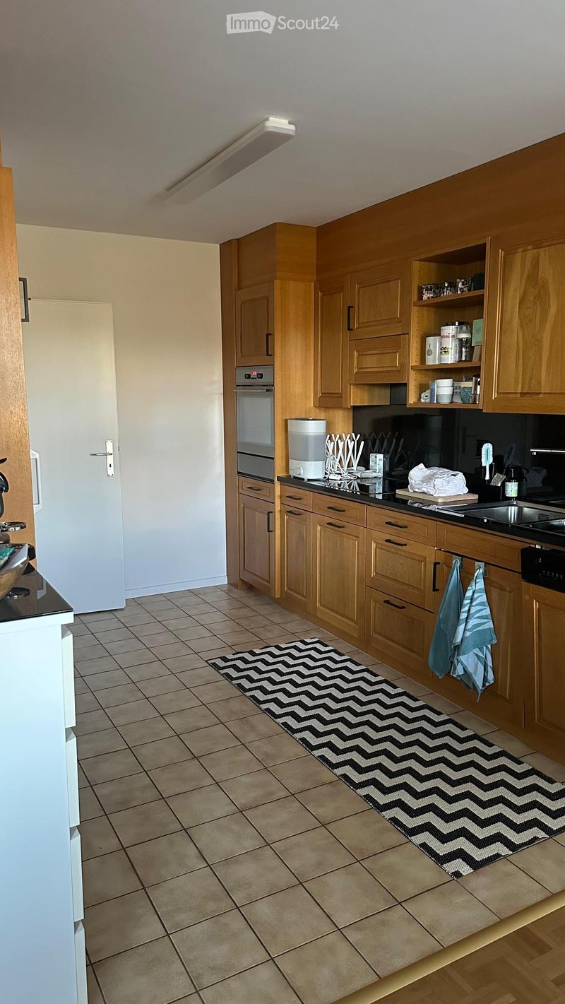 A modern kitchen with wooden cabinets, a tiled floor, and a black and white chevron-patterned rug. Visible appliances include a refrigerator, oven, and water dispenser. A cutting board and towels are on the counter.