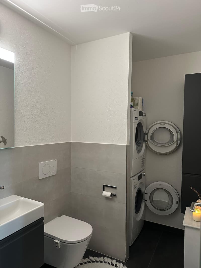 Modern bathroom with a sink, toilet, washing machine, dryer, and a small candle on a shelf.
