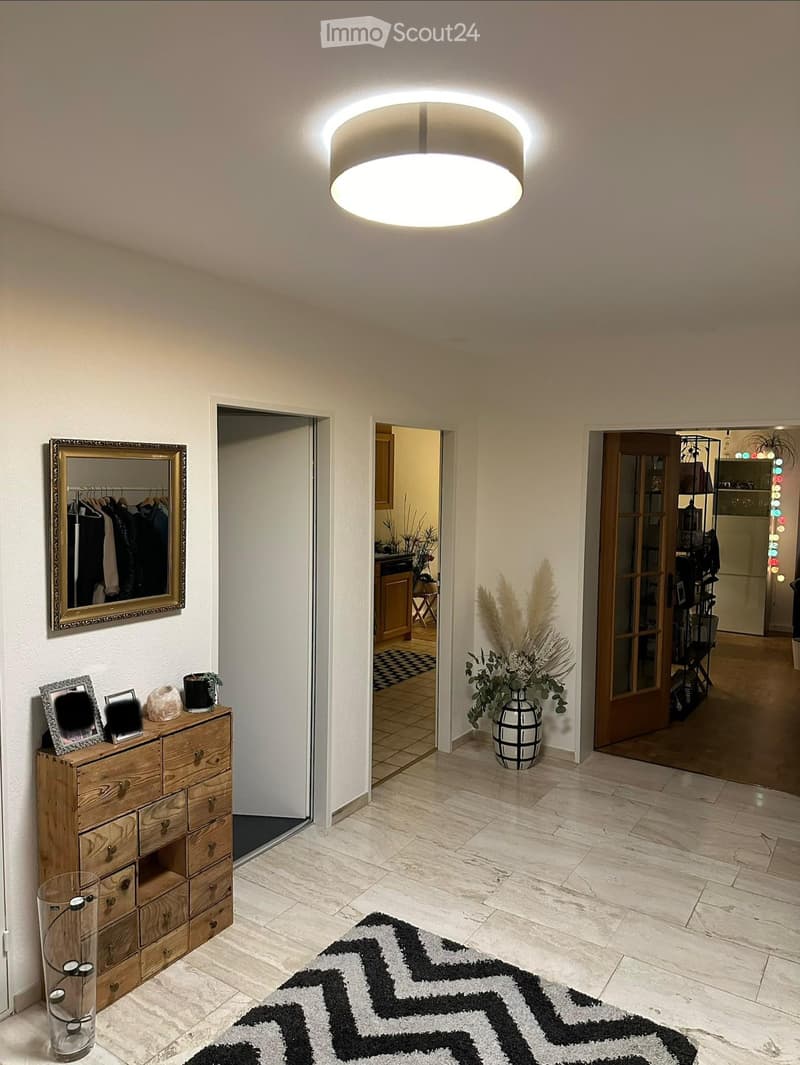 Bright hallway with marble flooring, modern ceiling light, wooden dresser with framed photos, mirror, and decorative vase with dried plants. Open doors lead to adjacent rooms.