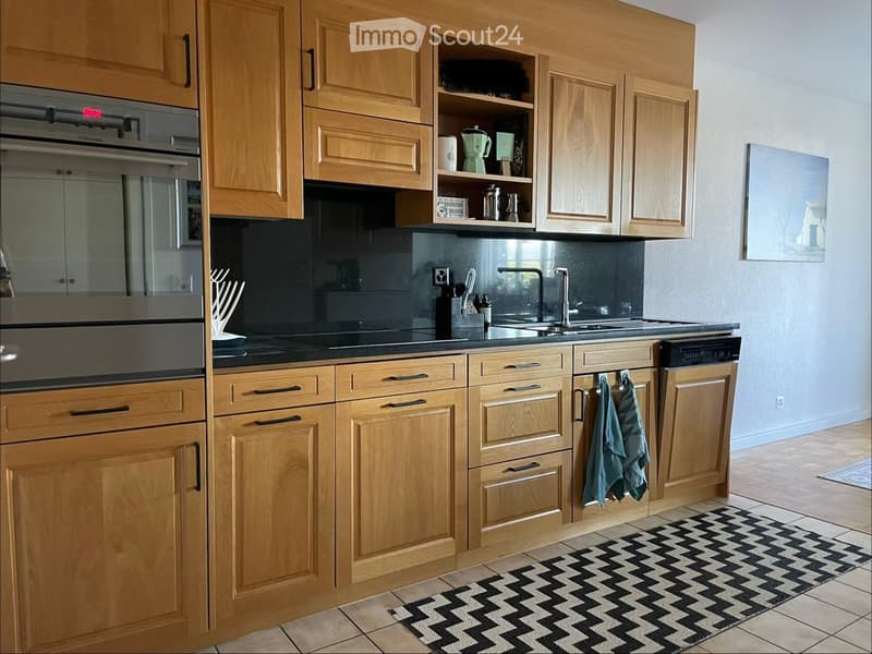 A modern kitchen with wooden cabinets, stainless steel appliances, a black countertop, and a black and white chevron-patterned rug.