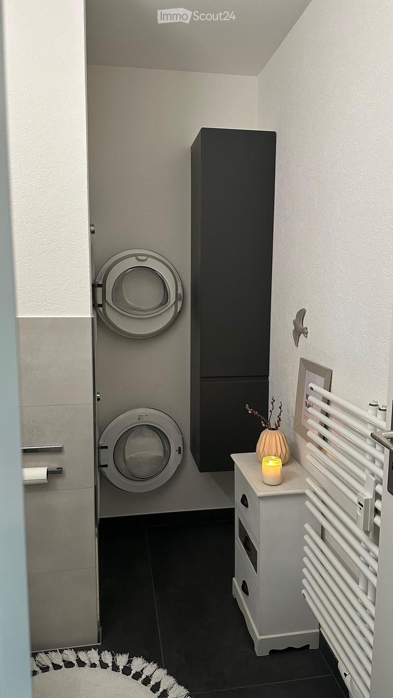 Modern bathroom with a built-in washing machine, radiator, candle, vase, and picture frame on a small cabinet.