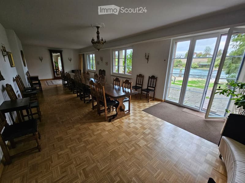 Spacious dining room with wooden herringbone flooring, large dining table surrounded by chairs, a sofa, a mirror, wall sconces, and a chandelier. Large windows and sliding glass doors lead to an outdoor area.