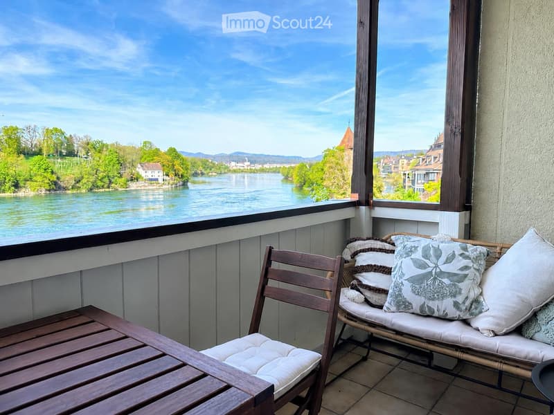 Balcony with wooden chair, bench, small table, and decorative pillows, overlooking a river and town.
