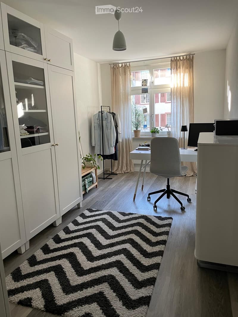 A modern, well-lit room with a desk, chair, cabinet, and a patterned rug. The room features large windows with curtains and a coat rack with jackets.