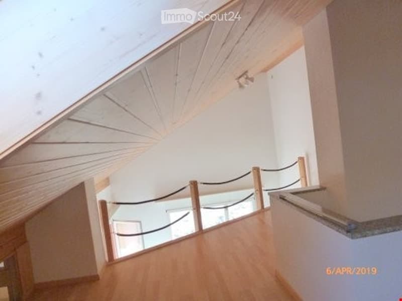 Spacious, modern hallway with wooden flooring, white walls, and a sleek glass railing leading to an upper level. The ceiling features exposed wooden beams and a recessed light fixture.