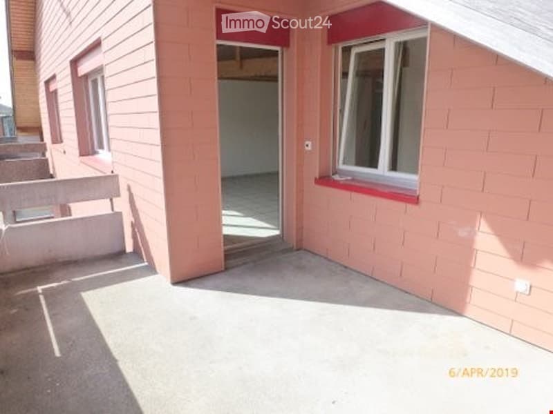 Empty balcony area with a view of an adjacent room. The balcony has a concrete floor, pink brick walls, and a white railing. The adjacent room is visible through an open doorway and appears to be unfinished with exposed walls and a concrete floor.