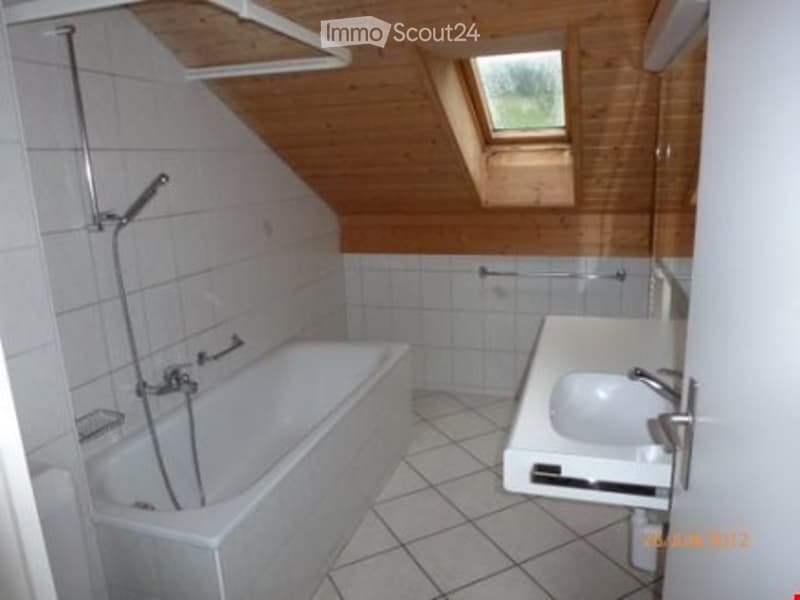 Bathroom with a bathtub, sink, and shower hose. The room features white tiled walls and floor, wooden ceiling beams, and a skylight window.