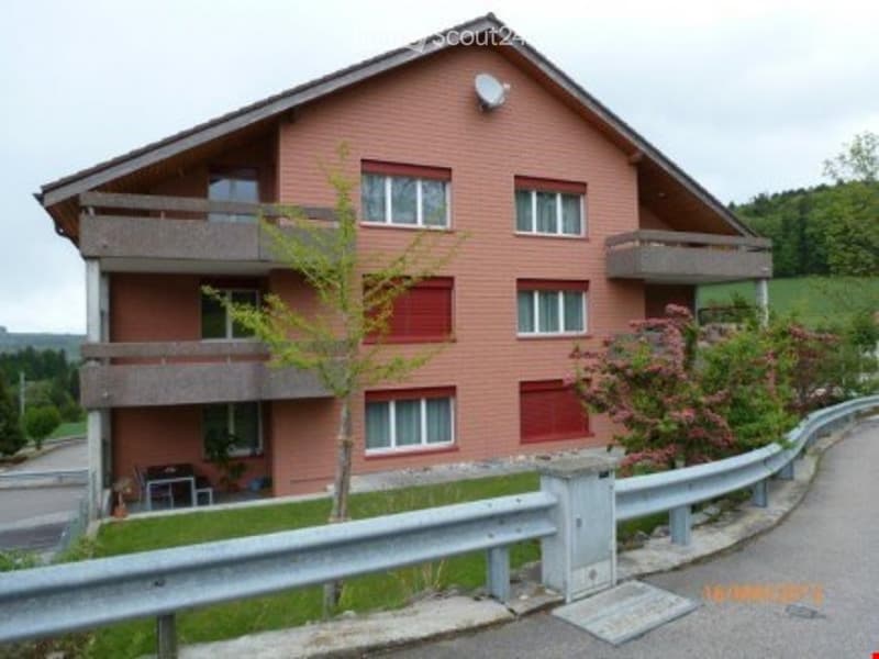 Two-story house with red brick facade, multiple windows, balconies, and a small garden area with a bench and a tree.