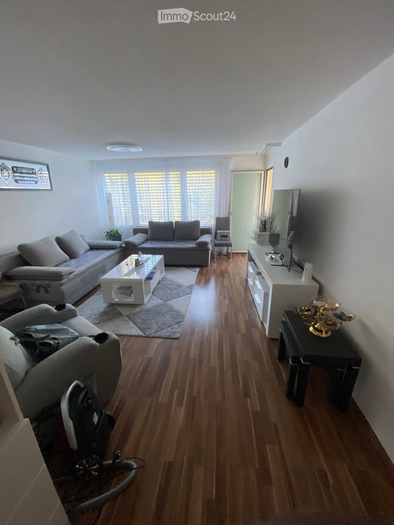 Modern living room with wooden flooring, large windows, grey sofa, coffee table, TV stand, and decorative items.