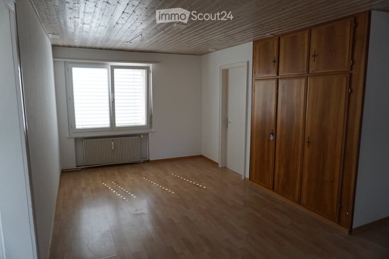 Empty room with wooden flooring, white walls, large window with blinds, radiator, and built-in wooden wardrobe.