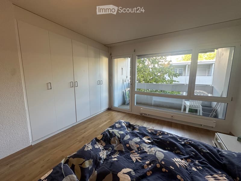 Bright bedroom with wooden flooring, large window opening to a balcony, bed with patterned bedding, and white built-in wardrobe.