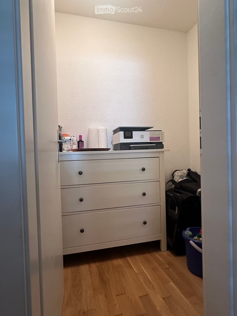 A small, furnished room with a wooden floor, a white drawer unit, a printer on top, and a backpack leaning against the wall.