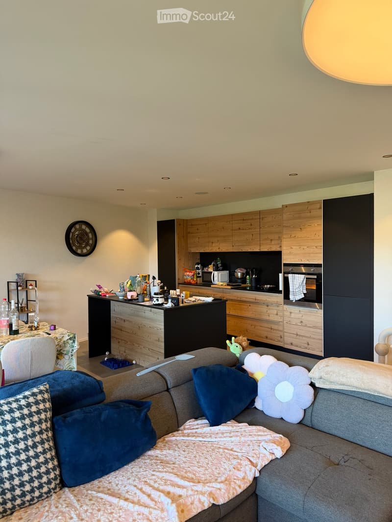 A modern, open-concept living and dining area with a grey sectional sofa, throw pillows, a coffee table, and a dining table with chairs. The kitchen features wooden cabinets, stainless steel appliances, and a microwave. A wall-mounted clock is visible on