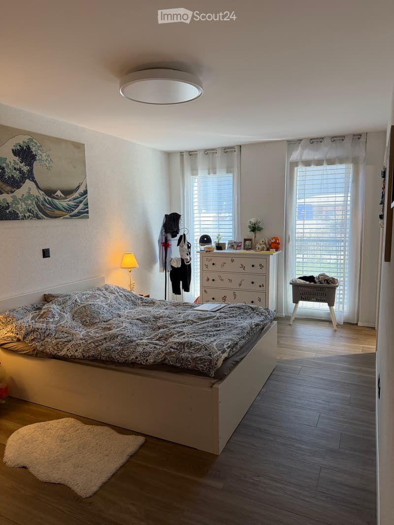 Cozy bedroom with a large bed, nightstand, lamp, and a drawer unit. Features a large window with blinds, a coat rack, and a decorative picture on the wall.