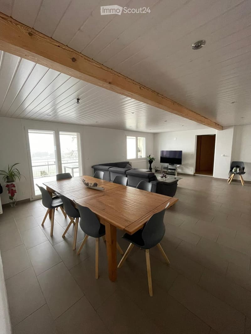 Bright, open living and dining area with wooden dining table, chairs, grey sofa, TV, and large windows leading to a balcony.