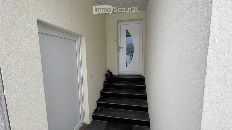 A short hallway with a raised ground floor leading to a white front door with a decorative glass panel. The walls are light-colored, and there is a small decorative item near the base of the stairs.
